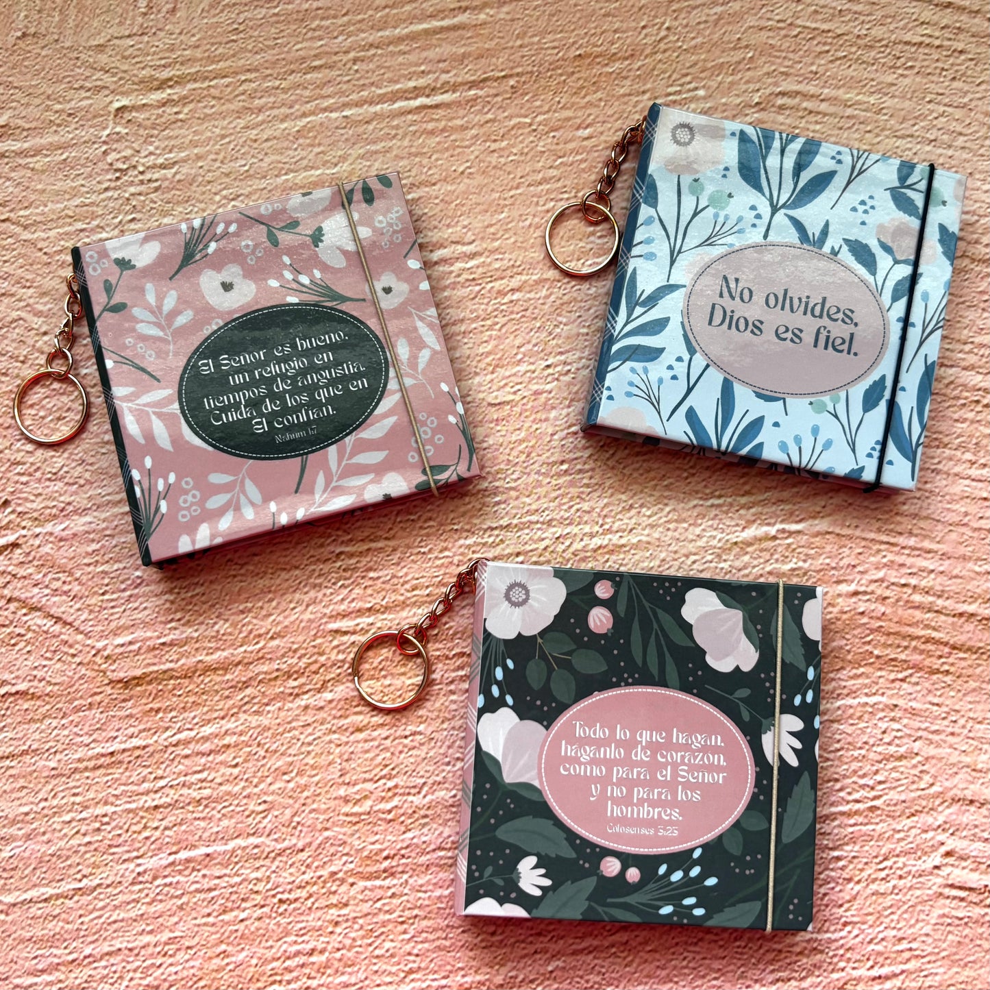 Three small floral keychains or bookmarks with Spanish text are arranged on a textured peach surface. One has a pink floral cover with a dark green oval label reading 'Saber el Camino es confiar'; one has a light blue floral cover with a beige oval label reading 'No olvides, Dios es fiel'; and one has a dark green floral cover with a pink oval label reading 'Boda la que hágan, legado de servicio como parte de Señor y de ellos, los hombres'.